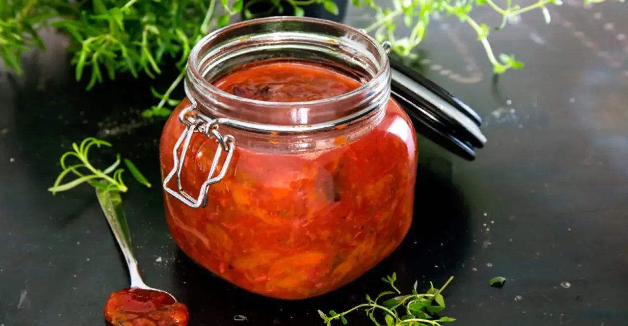 En burk med plommonchutney, perfekt för ett plommon recept. En sked med chutney ligger bredvid. Gröna örter ramar in scenen.