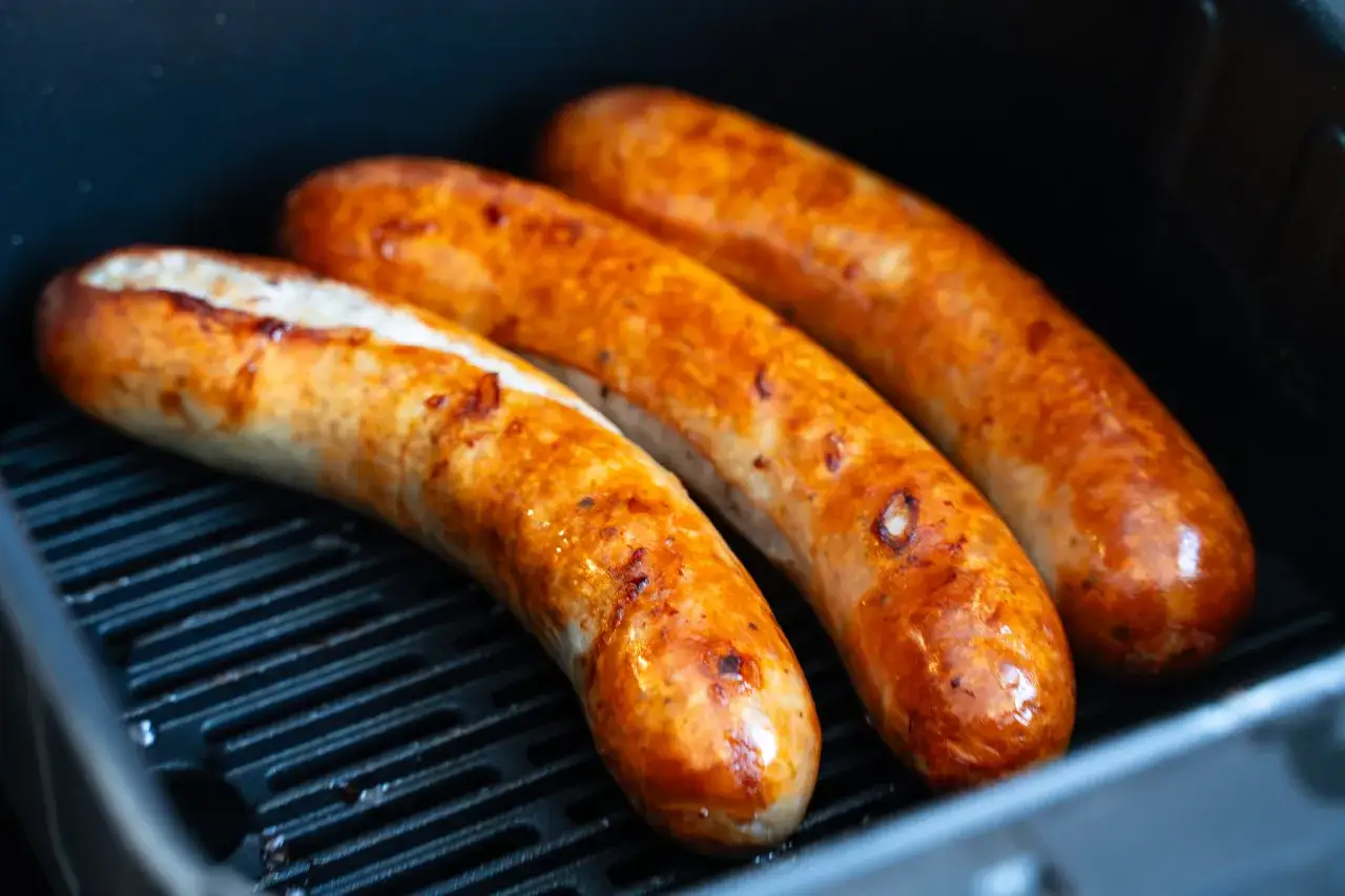 Drei goldbraune Bratw&uuml;rste liegen in einem Airfryer und werden knusprig.