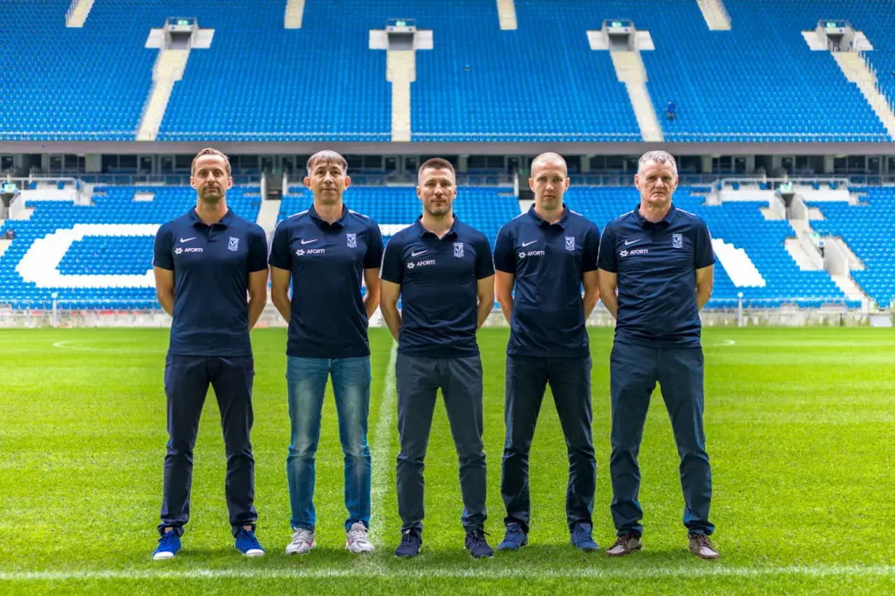 Pięciu mężczyzn w granatowych polo z herbem Lecha Poznań stoi na murawie stadionu. To lech poznan kadra, gotowa do walki.