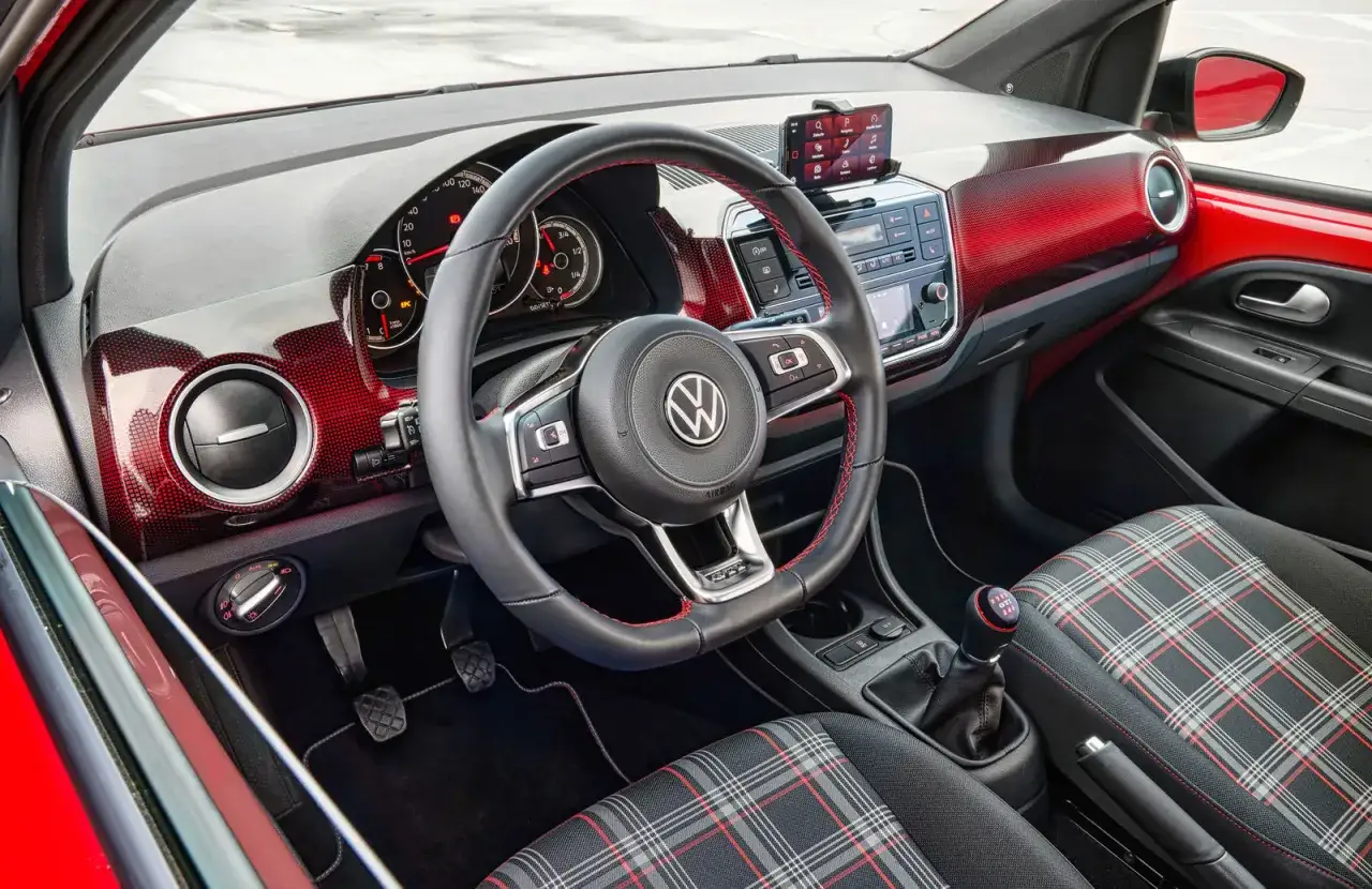 Interiör i en röd VW Up GTI med sportiga rutiga säten, en läderklädd ratt med röda sömmar och en växelspak med GTI-märkning.
