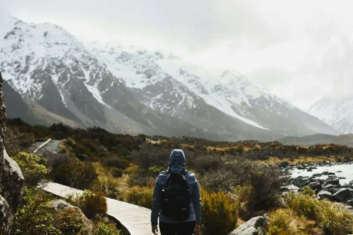 Une personne avec un sac à dos fait une randonnée sous la pluie, face à des montagnes enneigées. Le chemin en bois serpente à travers une végétation basse.
