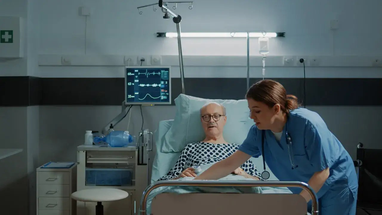 Opiekunka medyczna pochyla się nad pacjentem w szpitalnym ł&oacute;żku. To obraz z kursu opiekun medyczny.