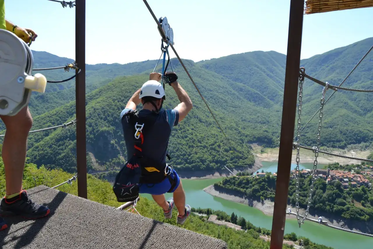 Un uomo in imbracatura si lancia in zip-line sopra il lago di Vagli, con montagne verdi e un borgo sottostante.