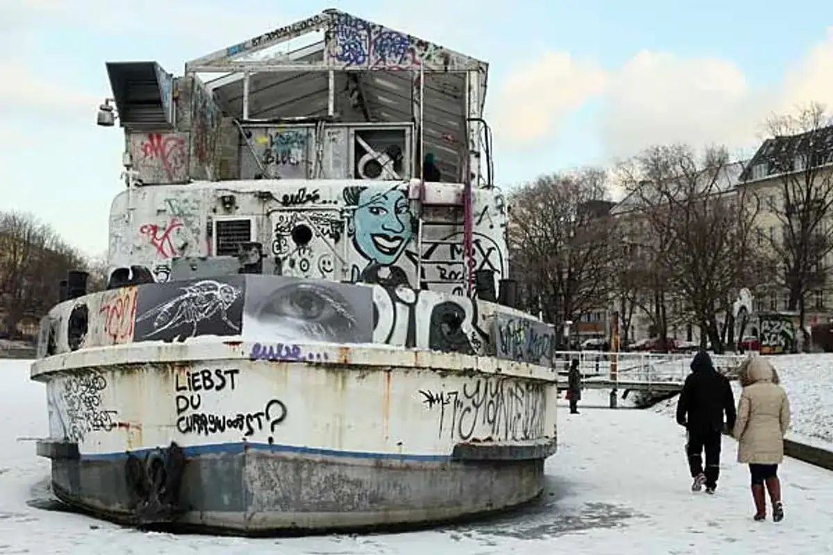 Graffiti-bespr&uuml;htes Schiff im Winter am Landwehrkanal Berlin. Ein Auge und eine Fliege zieren die Seite, w&auml;hrend zwei Personen im Schnee vorbeigehen.