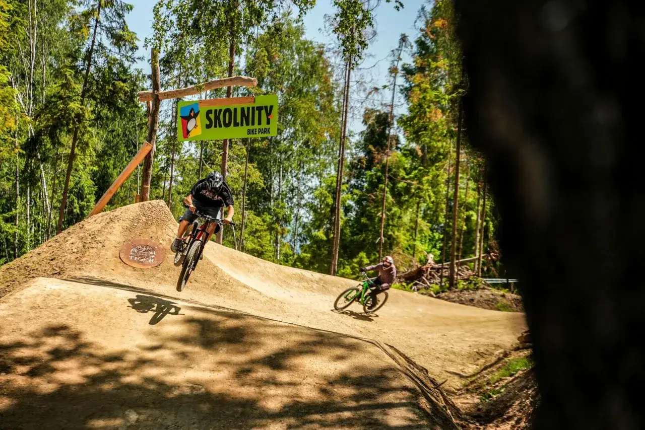 Dwóch rowerzystów zjeżdża po ziemnych **biking trails** w Skolnity Bike Park. Słońce przebija się przez drzewa.