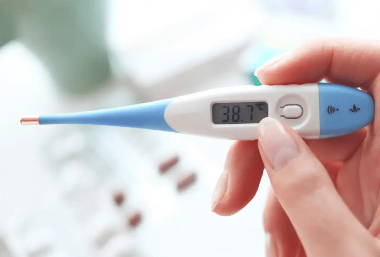 Thermomètre affichant 38.7°C. Une main tient l'appareil, suggérant la recherche de solutions pour comment faire descendre la fièvre adulte.