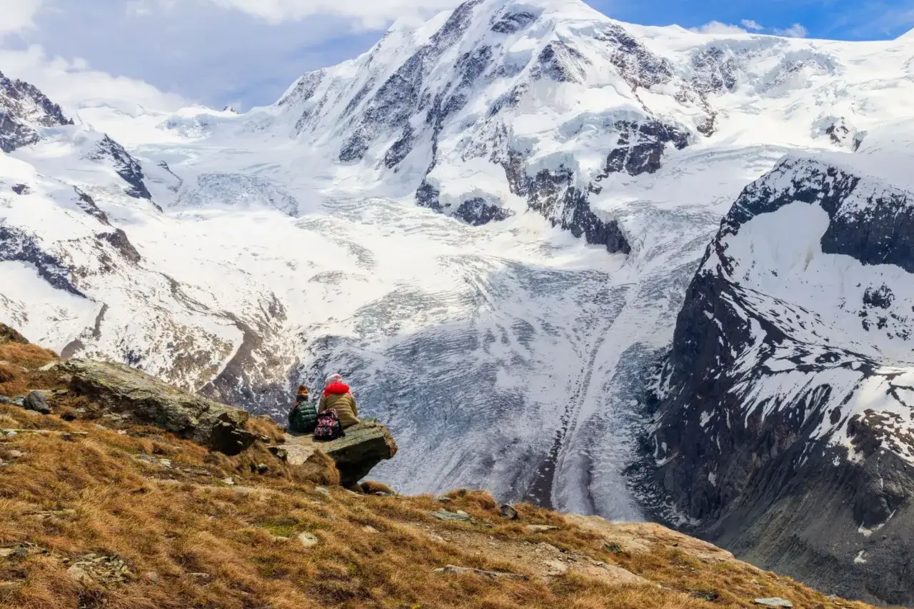 Two hikers rest on a rocky outcrop, gazing at the majestic, snow-covered peaks of the Tour de Monte Rosa.