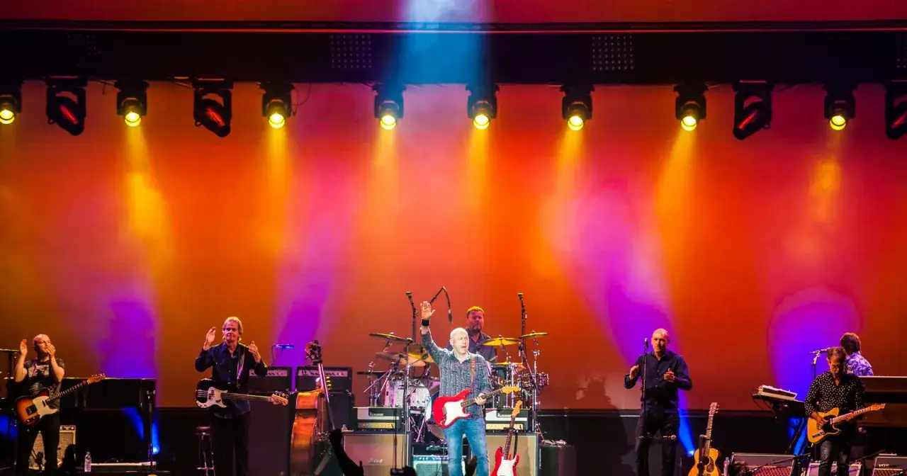 Zespół na scenie, grający utwory Dire Straits. Wokalista z gitarą, muzycy z basem i perkusją, w blasku żółtych i pomarańczowych świateł.