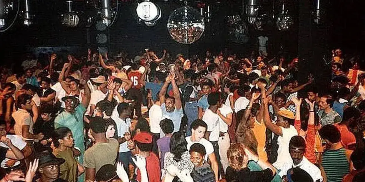 Una multitud baila en una discoteca, con luces de bola de espejos y un ambiente vibrante, evocando el esp&iacute;ritu del house, el g&eacute;nero musical.