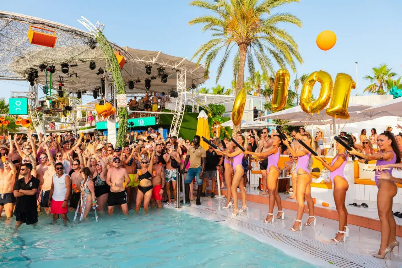 Tłum szaleje na basenie, świętując ibiza imprezy. Dziewczyny w fioletowych strojach wystrzeliwują konfetti, a nad nimi unoszą się złote balony z cyfrą 