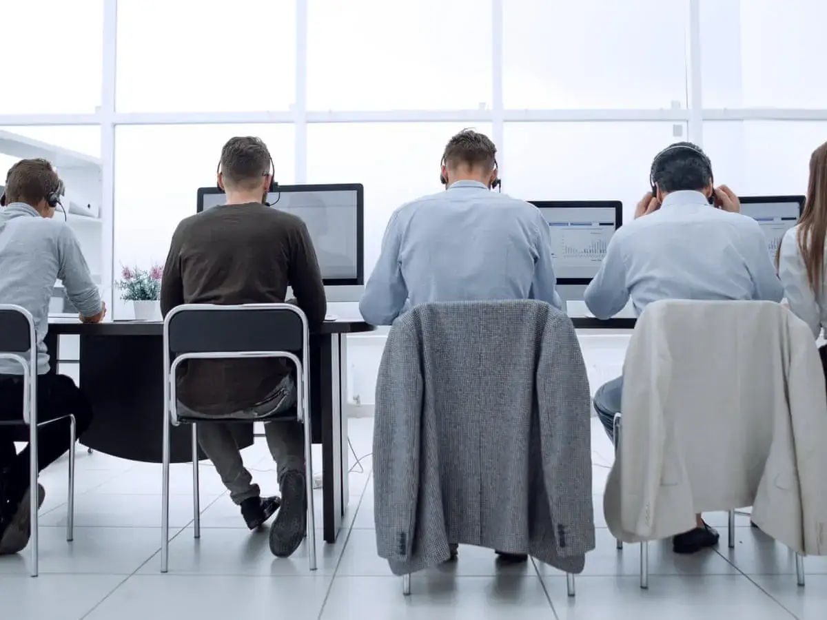 Call center employees, heads bowed, appear demoralized at work. Their backs are to the camera as they stare at computer screens.