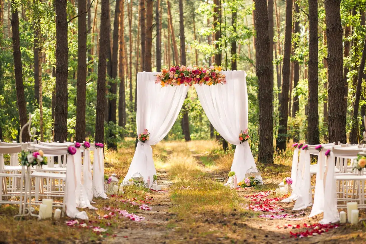 Un chemin de pétales mène à une arche fleurie, décorée pour un mariage champêtre. Des chaises blanches ornent la forêt.