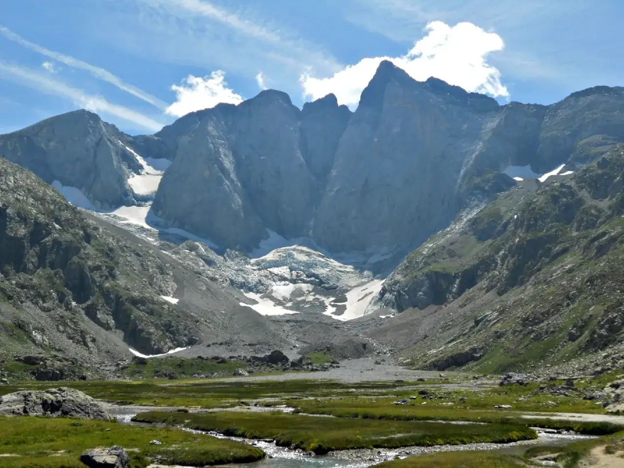 Majestuosas montañas y un glaciar en el refugio Oulettes de Gaube. Un arroyo serpentea por un prado verde.