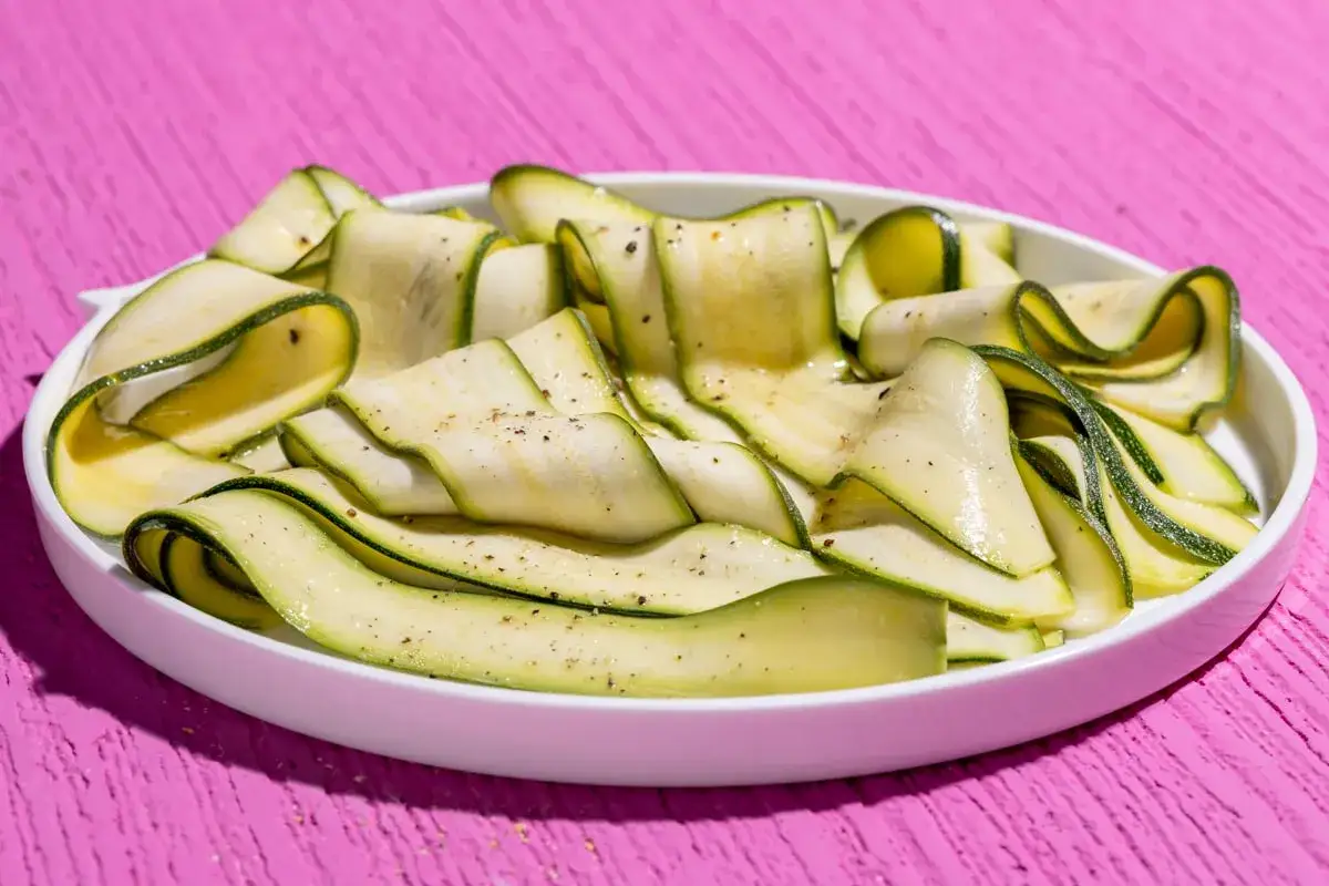 Fettine di zucchine marinate cotte, condite con pepe nero, disposte a spirale in un piatto bianco su un tavolo rosa acceso.