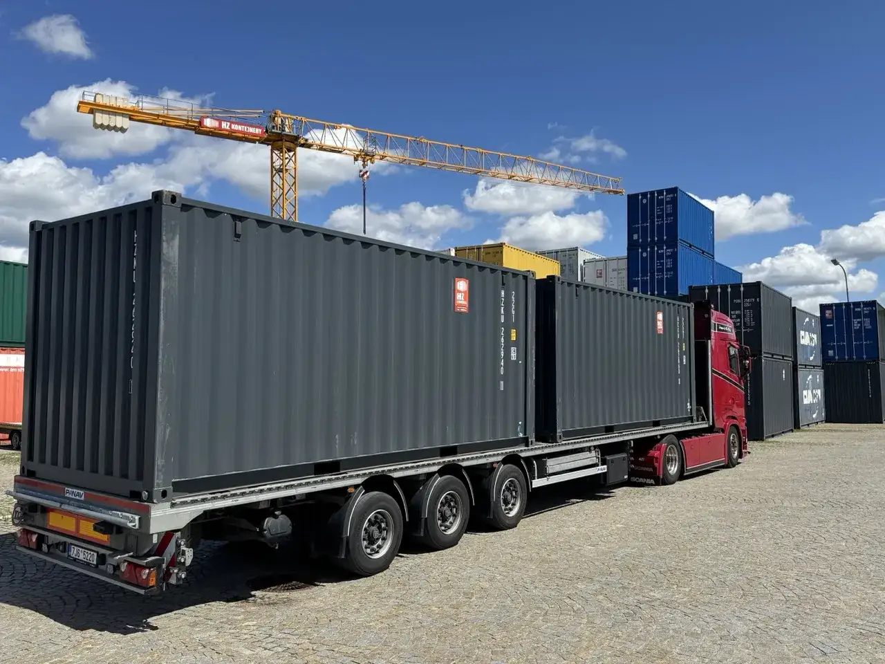 Ein roter LKW mit zwei grauen Containern wartet auf seine Abfahrt. Im Hintergrund sind weitere Container und ein Kran zu sehen.