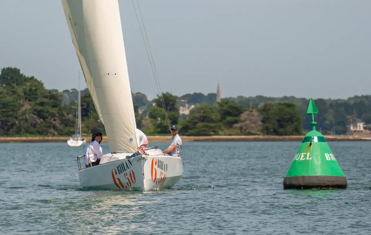 Un voilier BIHAN 6.50 navigue sur l'eau, deux personnes à bord. Une bouée verte est visible à droite.