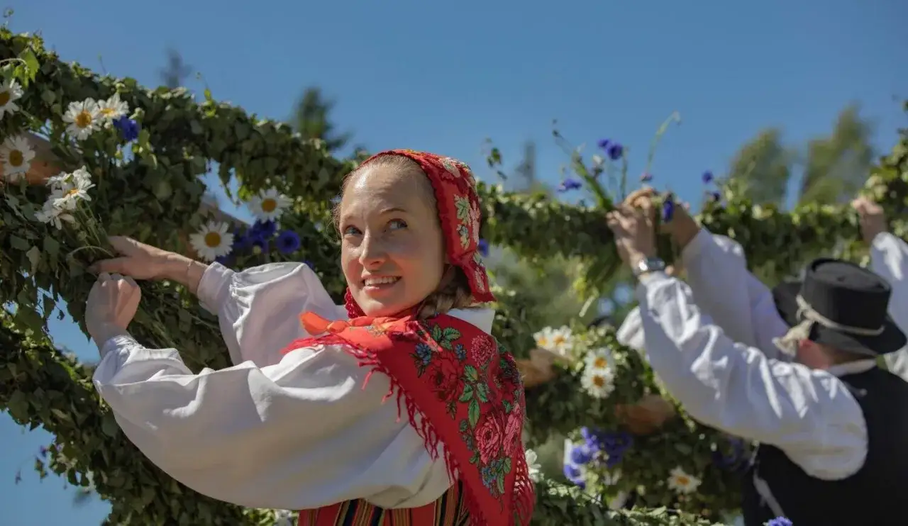 Glad kvinna i folkdräkt dekorerar midsommarstången på Skansen med blommor och grönt.