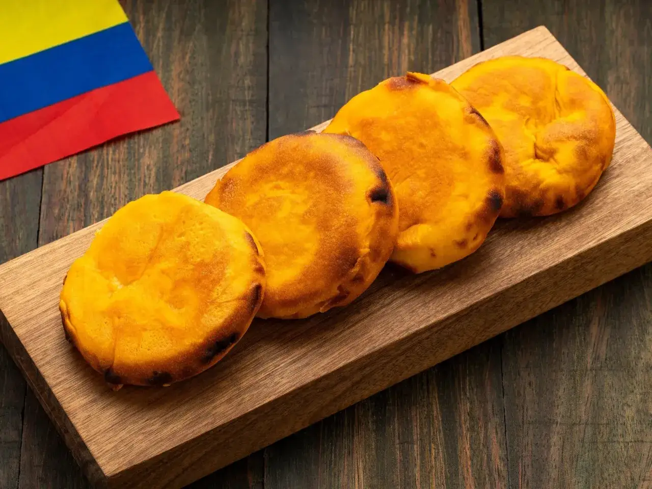 Cztery złociste arepas leżą na drewnianej desce, obok flagi Kolumbii.
