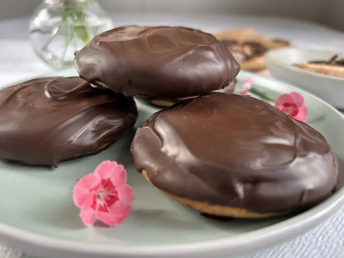 Tre chokladöverdragna biskvier på en ljusgrön tallrik, med små rosa blommor. Ett perfekt biskvier recept för fikat.