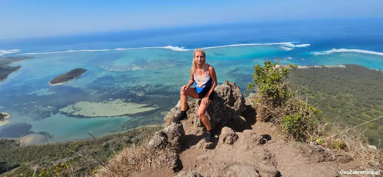 Kobieta siedzi na skale z widokiem na turkusową lagunę i ocean. W tle widać wyspę Ilot Sancho i zielone zbocza Le Morne Mauritius.