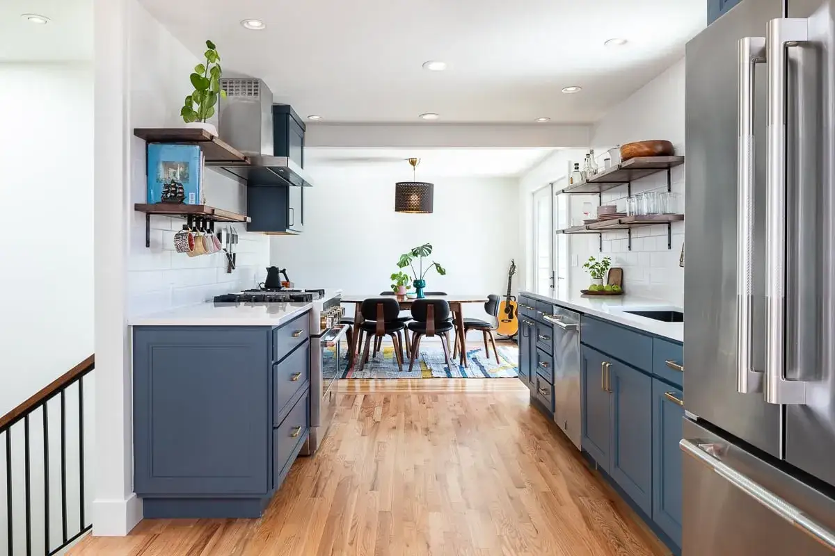A modern kitchen with deep blue cabinets, white subway tile backsplash, and stainless steel appliances. The warm wood floors and colorful rug add a cozy touch.