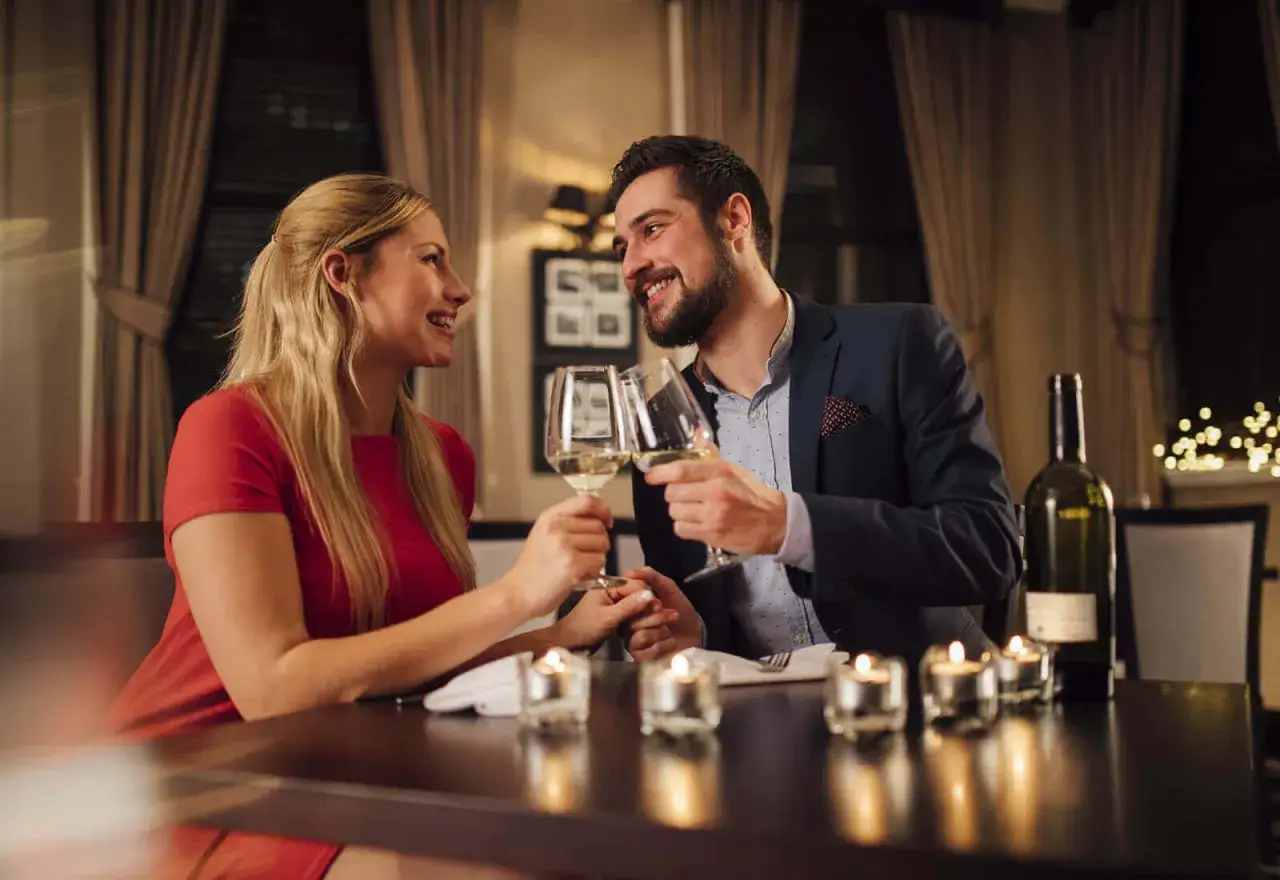 Pareja brindando con copas de vino blanco para celebrar aniversario en un restaurante rom&aacute;ntico, con velas y luces tenues.