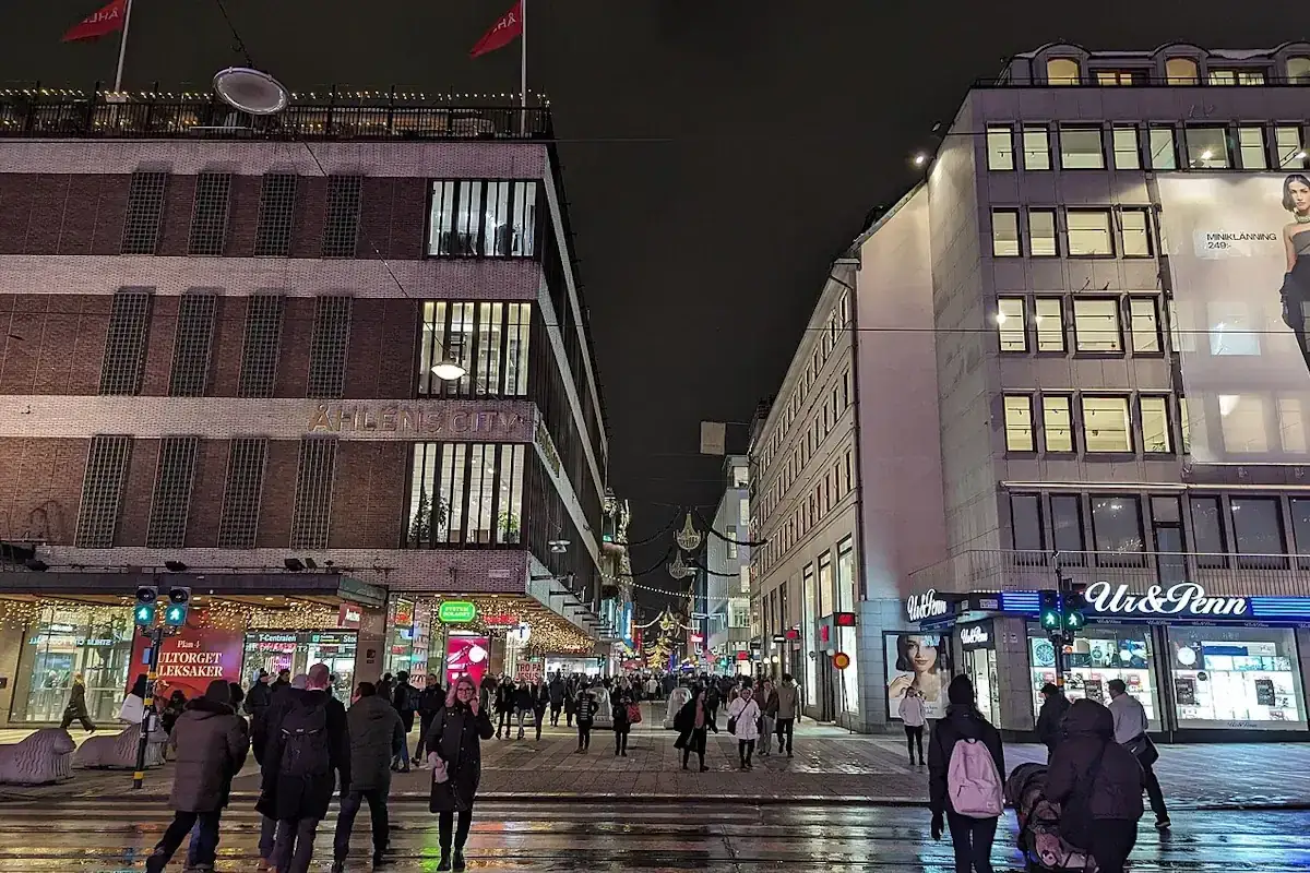 En livlig stockholm shopping street på kvällen. Folk promenerar förbi Åhléns City och Ur & Penn under juldekorationer.