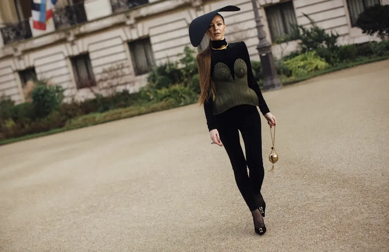 Une femme porte une tenue avec legging noir, un corset vert olive et un chapeau noir audacieux, tenant un sac dor&eacute;.