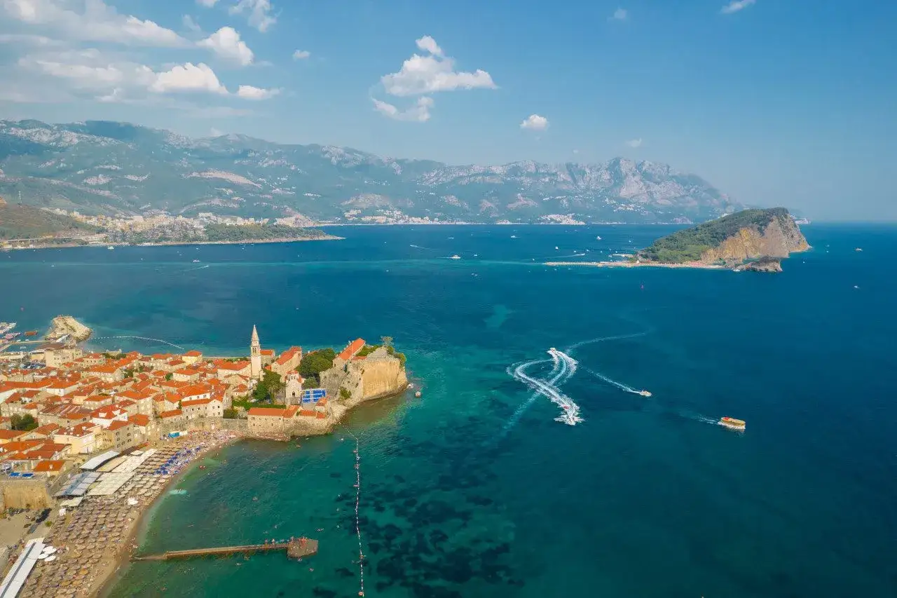 Widok z lotu ptaka na Budva stare miasto z czerwonymi dachami, plażą i turkusowym morzem. W oddali widać wyspę Sveti Nikola i góry.