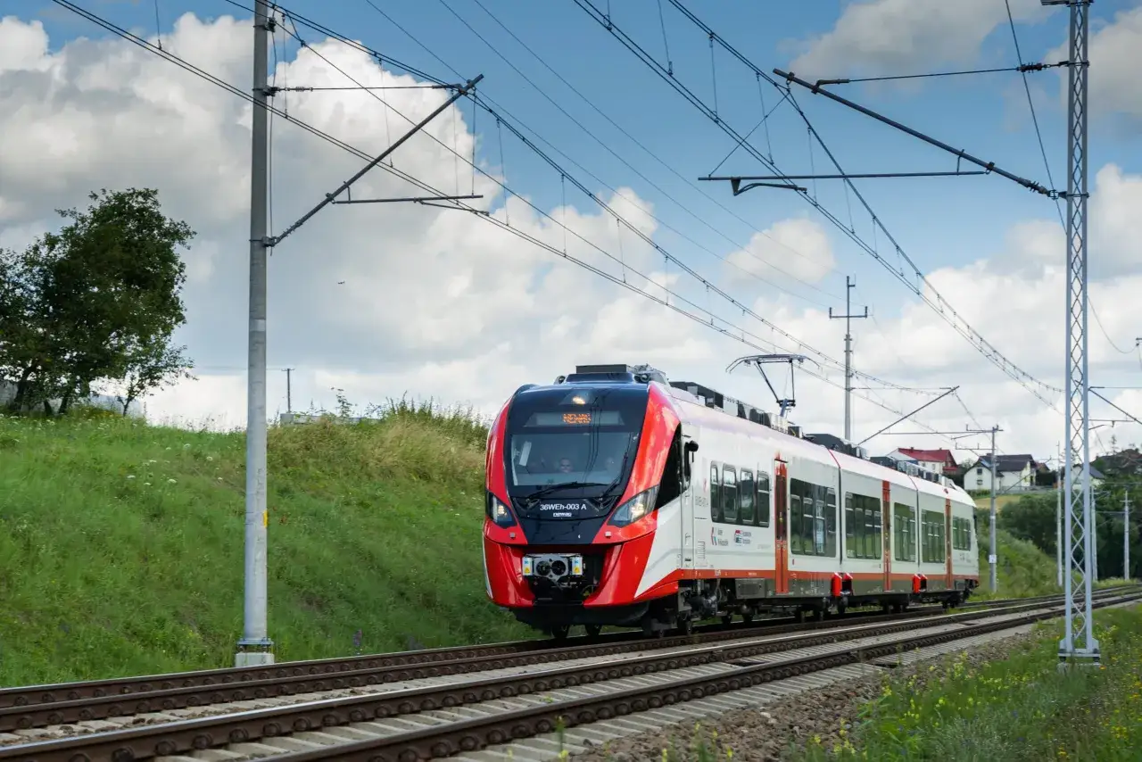 Nowoczesny autobus szynowy w czerwono-białych barwach jedzie po torach, mijając zielone wzgórza i domy.