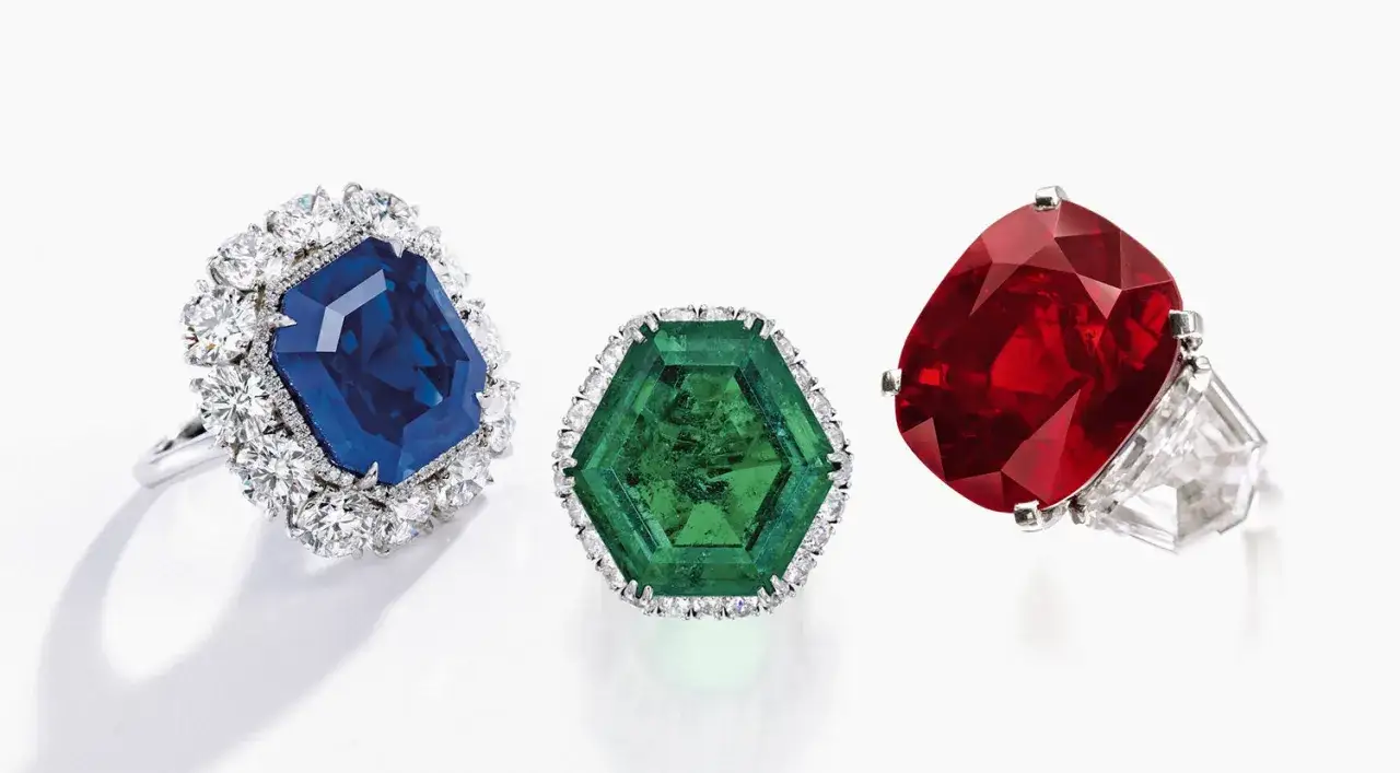 Tres anillos con deslumbrantes piedras preciosas: un zafiro azul rodeado de diamantes, una esmeralda hexagonal y un rubí rojo intenso.