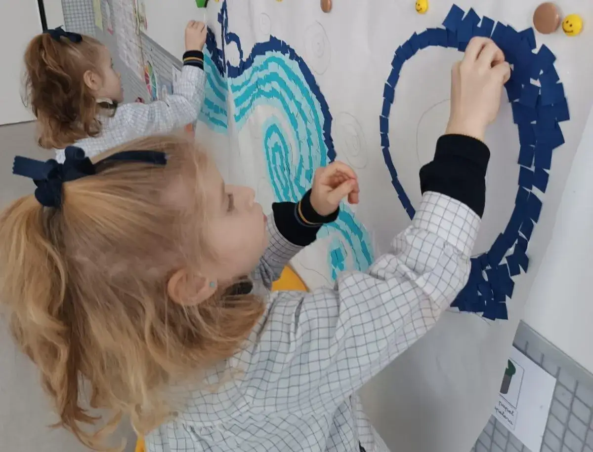 Niñas pegan trozos de papel azul en un mural, desarrollando habilidades clave para el perfil de salida LOMCE.