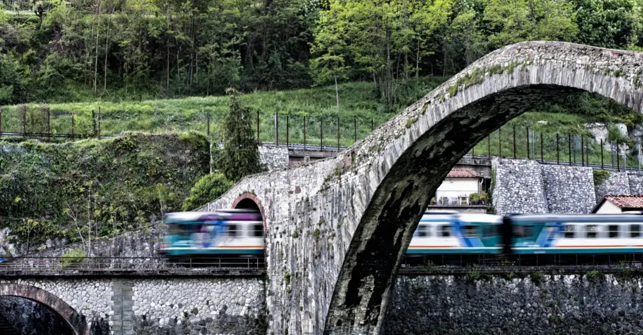Mit dem Zug in die Toskana: Ein Zug fährt über eine alte Steinbrücke, umgeben von üppigem Grün.