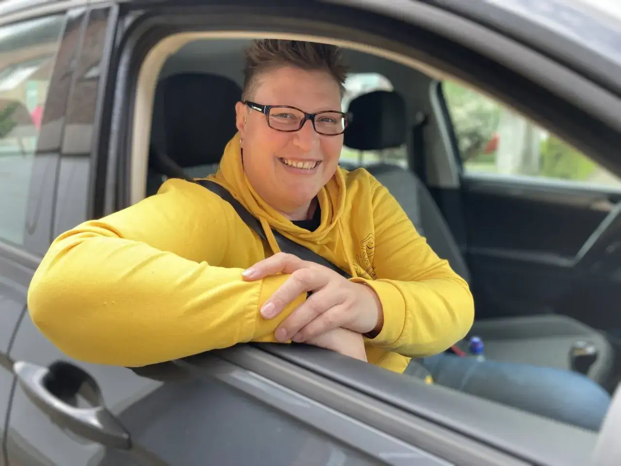 Lächelnde Person im gelben Hoodie lehnt am offenen Autofenster. Ein Symbol für die sichere Fahrtauglichkeit von Senioren, wie sie der ADAC unterstützt.