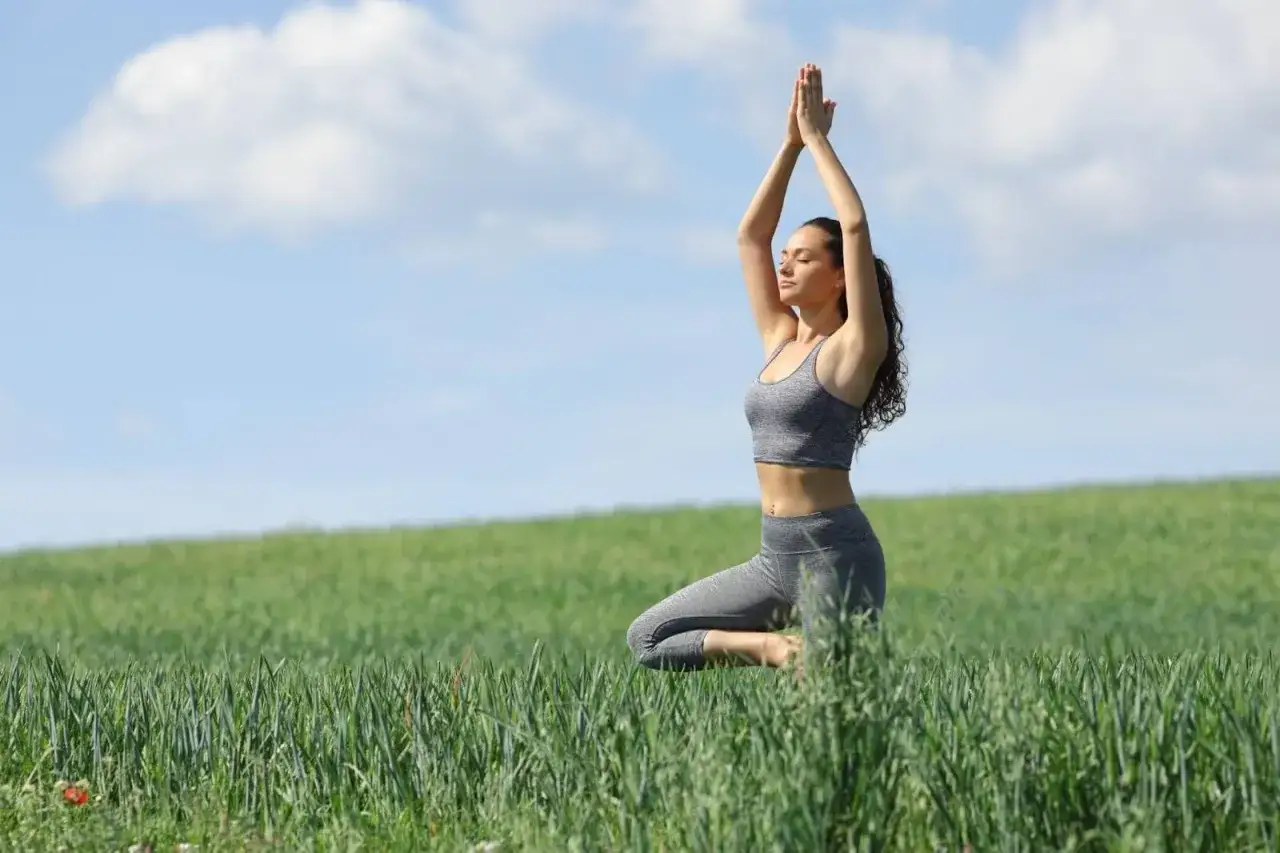 A woman practices one of 10 mindful exercises in a grassy field, balancing in a yoga pose with arms raised.