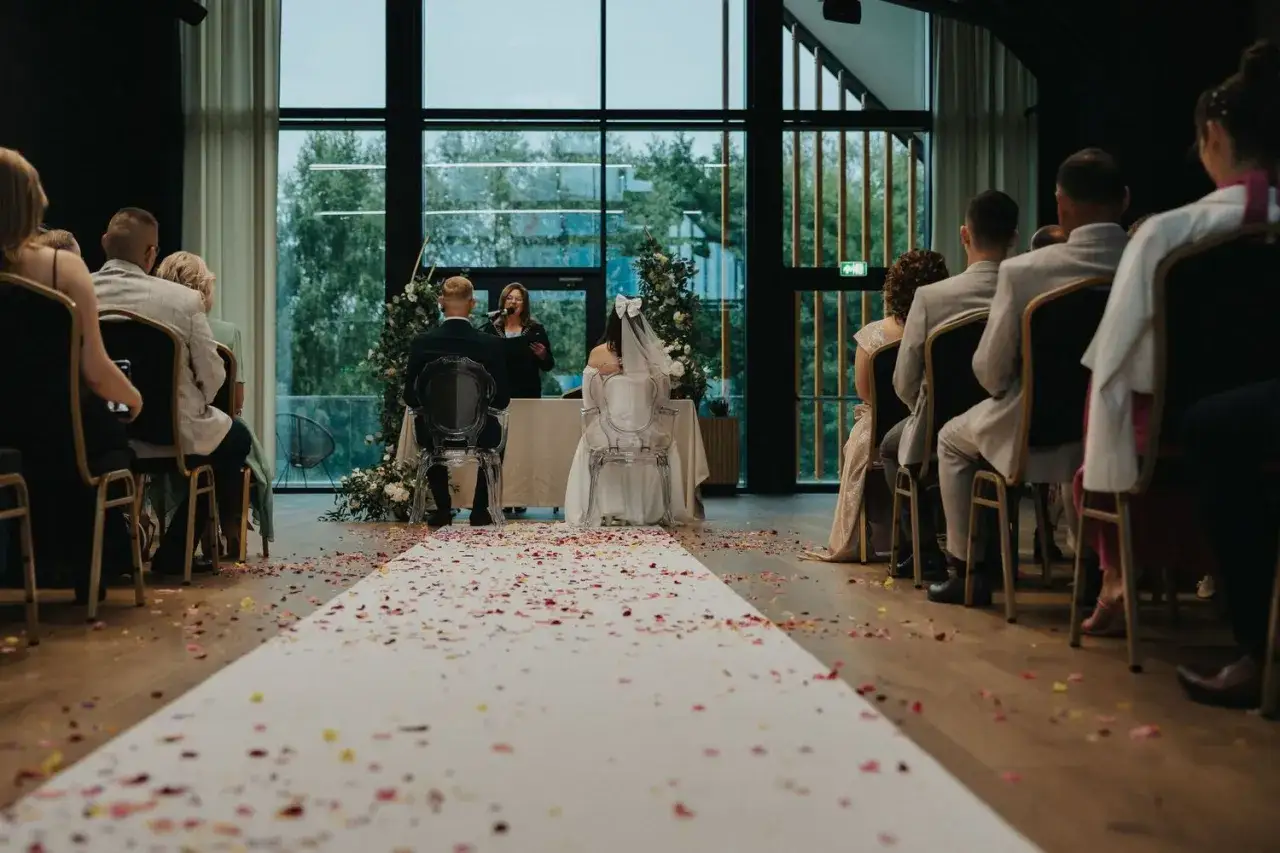 Para młoda siedzi przed urzędniczką podczas ceremonii ślubnej, na podłodze płatki róż.