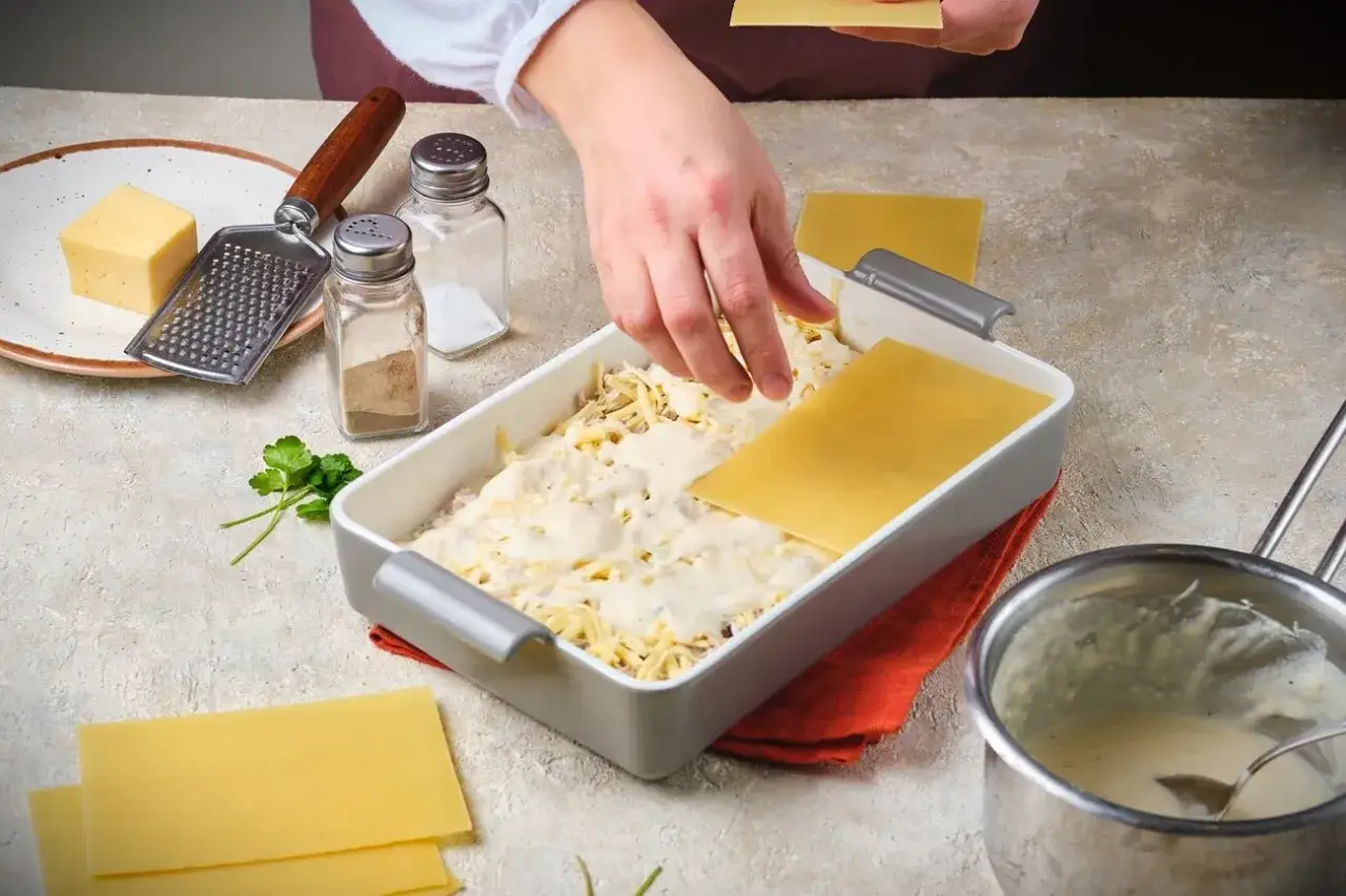 H&auml;nde legen Lasagneplatten auf eine cremige F&uuml;llung. Zutaten f&uuml;r eine vegetarische Lasagne liegen bereit.