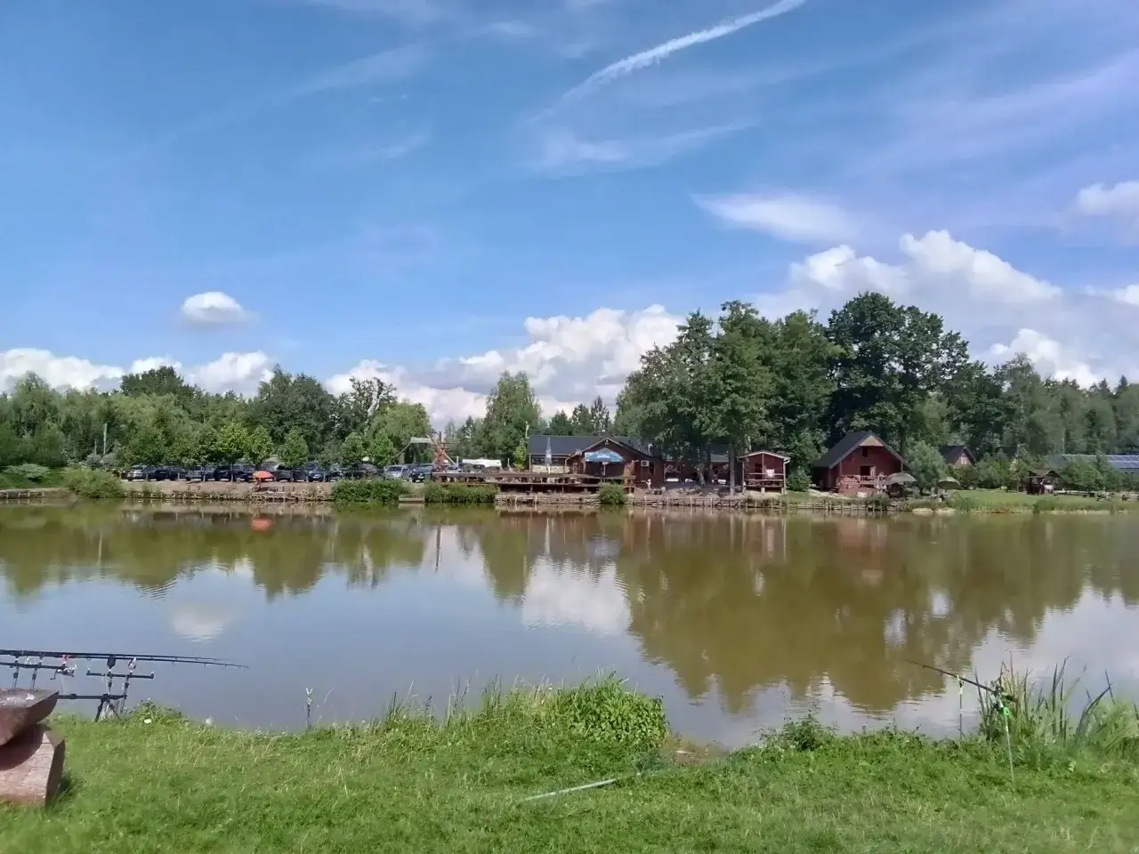 Idealna pogoda sielska woda, cisza i spokój nad jeziorem. Wędki czekają na branie, a nad brzegiem stoją urokliwe domki.
