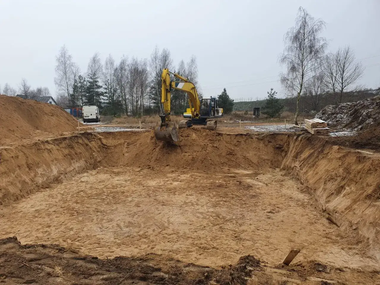 Koparka pracuje przy wykopy pod fundamenty. W tle biały van i stos desek.