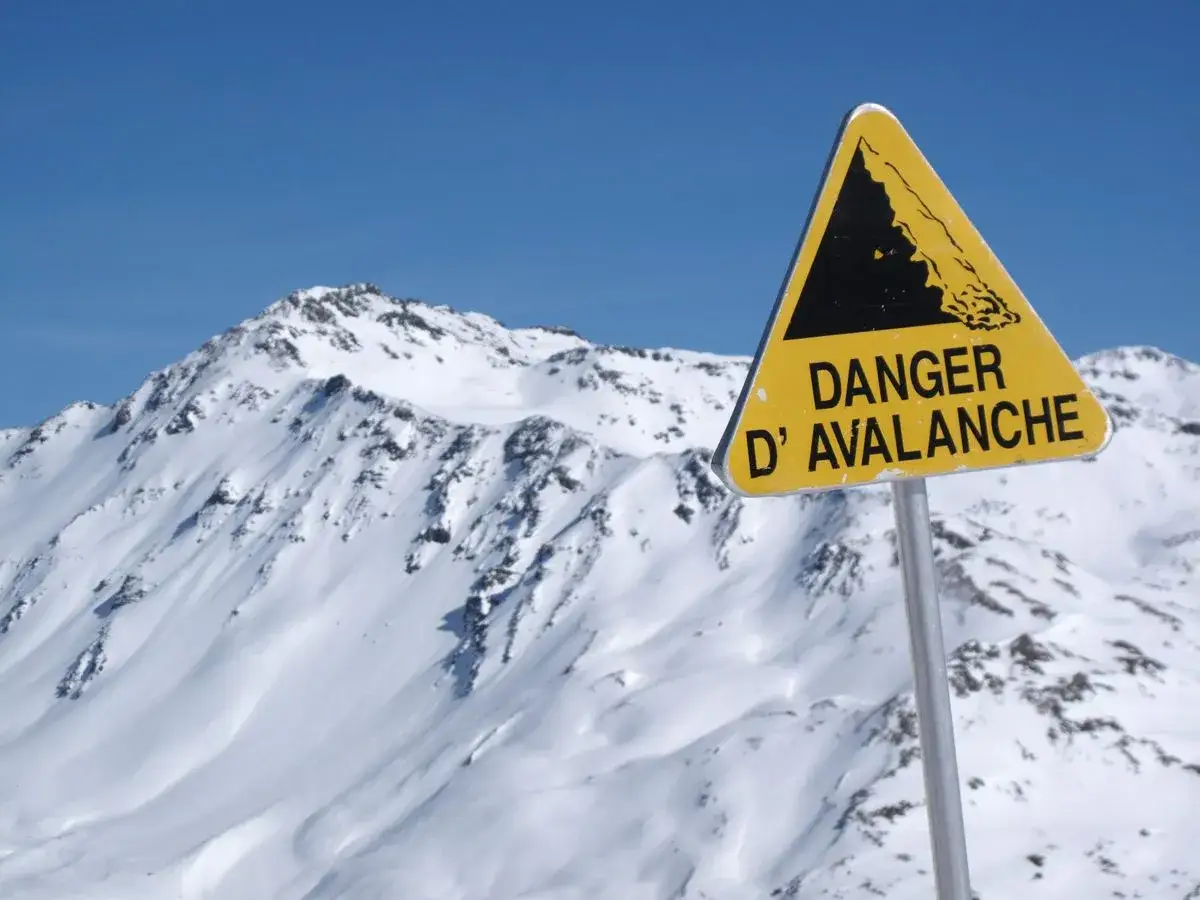 Panneau jaune triangulaire signalant le danger d'avalanche sur fond de montagnes enneigées et de ciel bleu. Les neiges mouvantes invitent à la prudence.