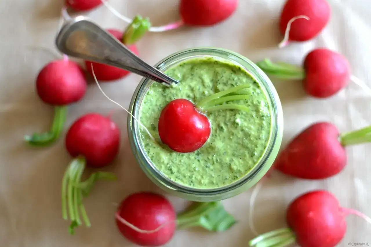 Pesto z liści rzodkiewki w słoiku, ozdobione rzodkiewką, otoczone świeżymi rzodkiewkami.
