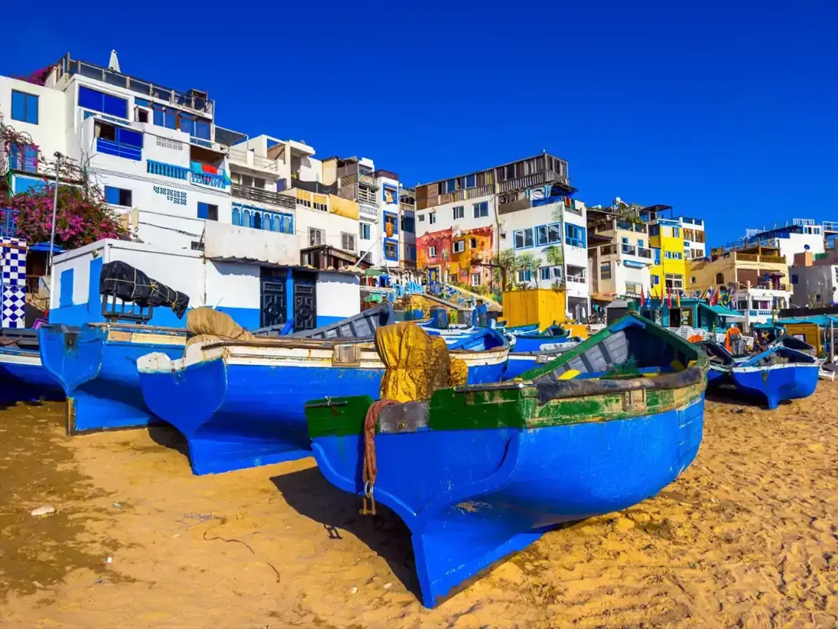 Blå fiskebåtar på sandstranden i Agadir, Marocko, med färgglada hus i bakgrunden under en klarblå himmel.