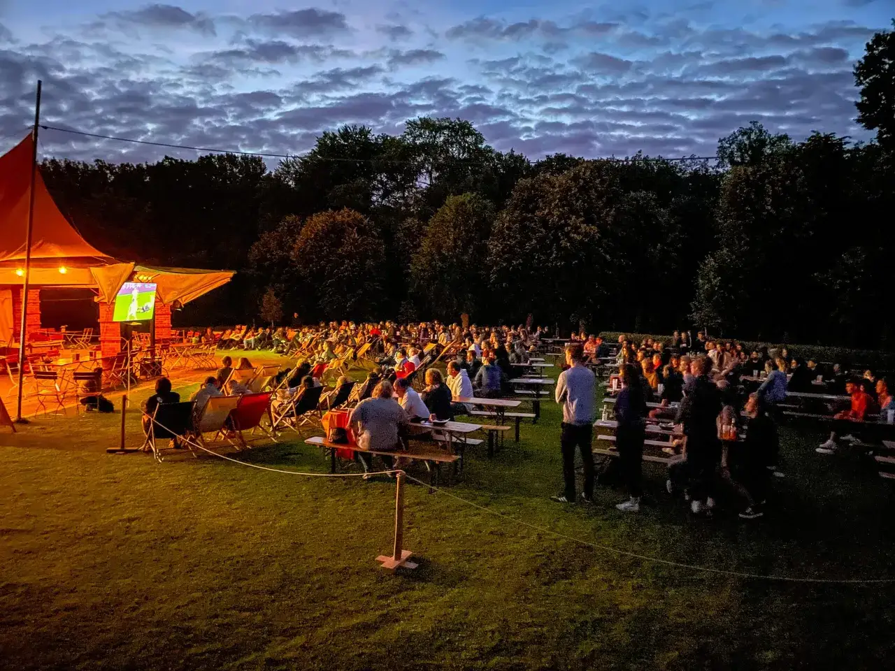 Menschen genie&szlig;en ein Fu&szlig;ballspiel bei einem **Public Viewing in Berlin**. Viele sitzen in Liegest&uuml;hlen und an Tischen im Freien, w&auml;hrend der Himmel sich verdunkelt.