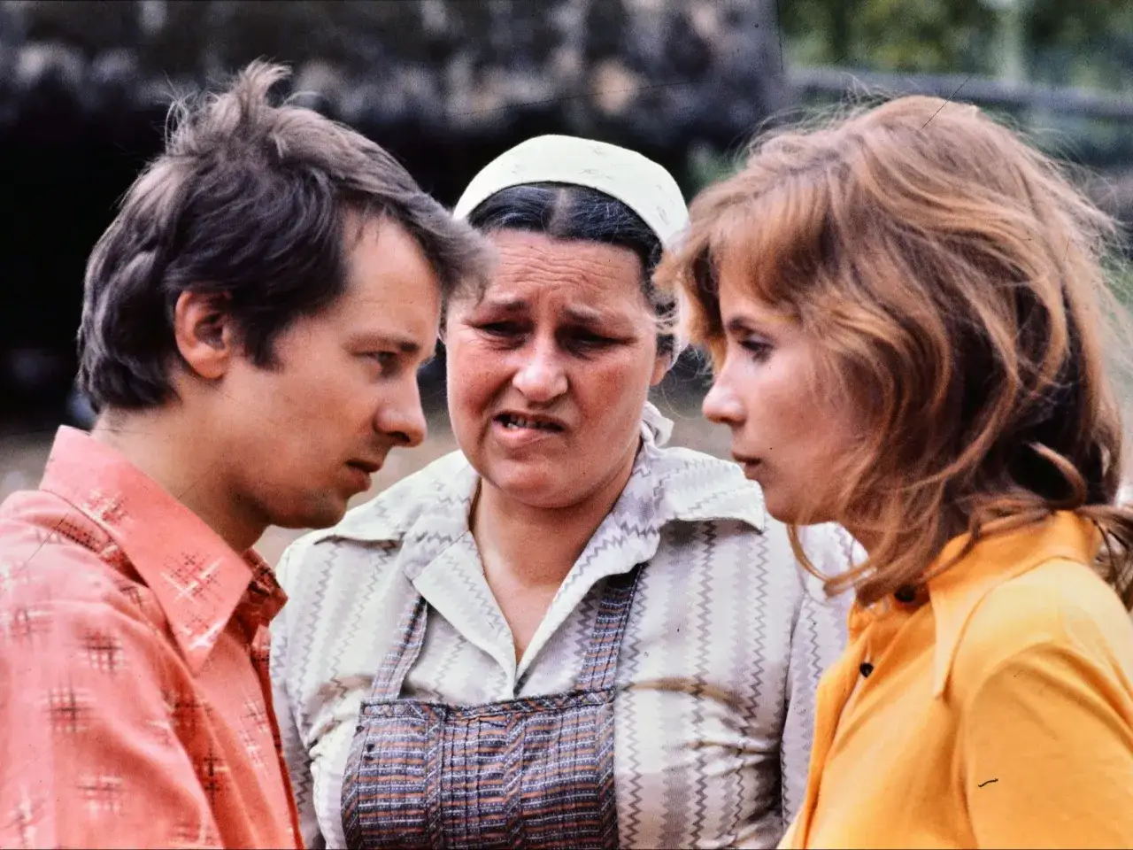 Trójka bohaterów, jak z kultowych polskich seriali, w rozmowie. Mężczyzna w różowej koszuli, kobieta w chustce i fartuchu, oraz kobieta w żółtej bluzce.