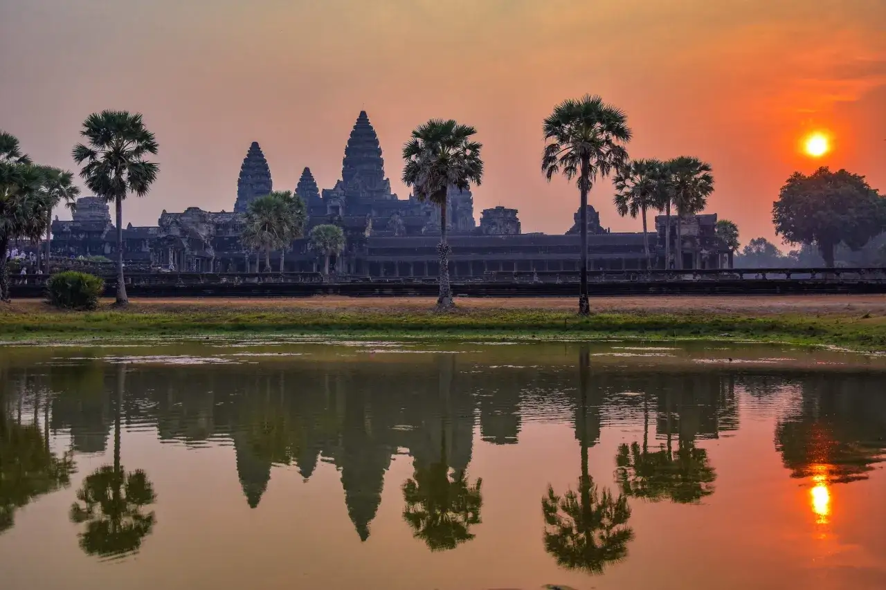 Zach&oacute;d słońca nad Angkor Wat, odbijający się w wodzie. Palmy i wieże świątyni tworzą magiczny widok. Oto angkor wat ciekawostki.