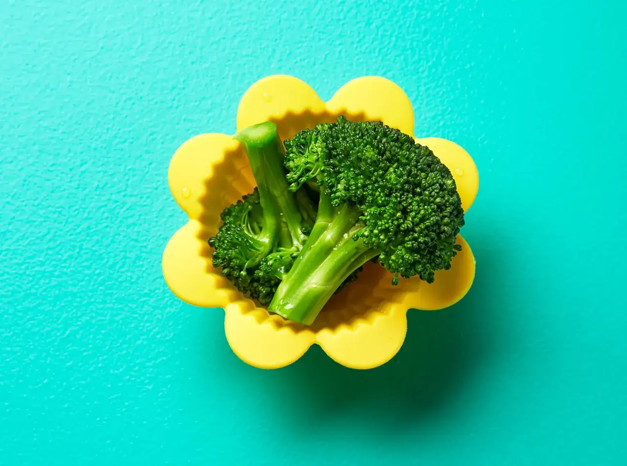 Frischer baby brokkoli in einer gelben Blumenform-Silikonform auf türkisfarbenem Hintergrund.