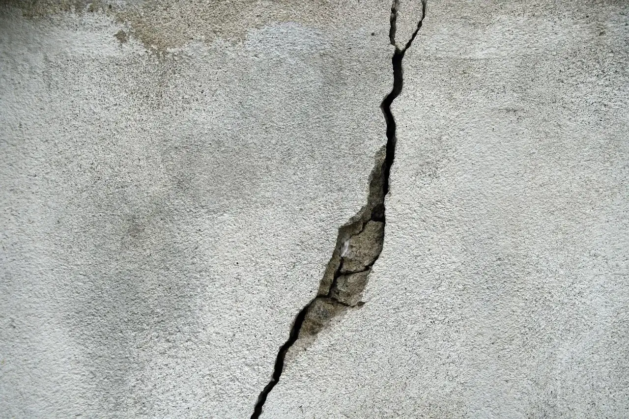 Szara ściana z widocznym pęknięciem, kt&oacute;re odsłania fragmenty betonu. To dopuszczalne rysy na tynku, świadczące o jego wieku.