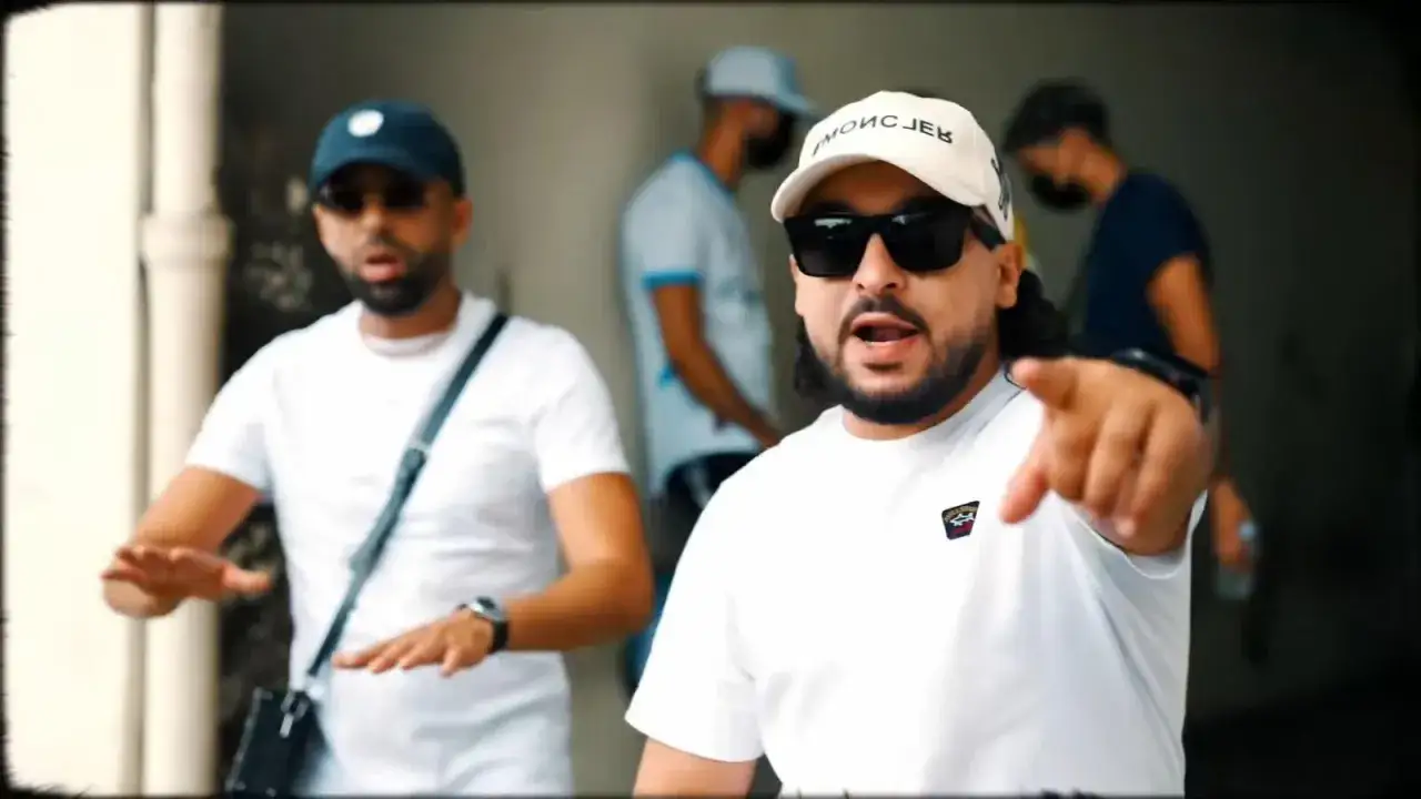 Deux rappeurs, symboles du hip hop en France, font un geste vers la caméra. L'un porte une casquette blanche, l'autre une casquette bleue.