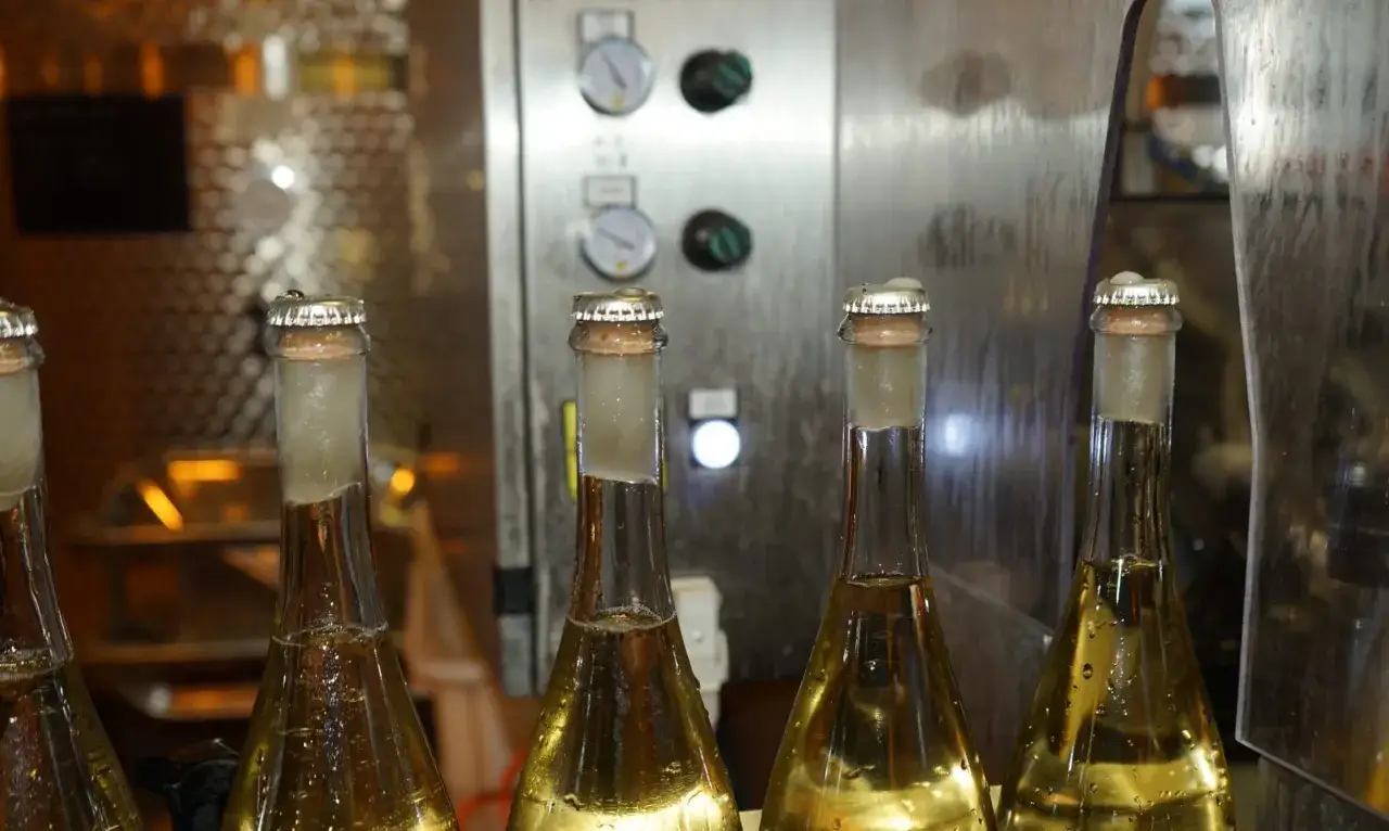 Bottiglie di vino pronte per la sboccatura, con tappi a corona e un macchinario industriale sullo sfondo.