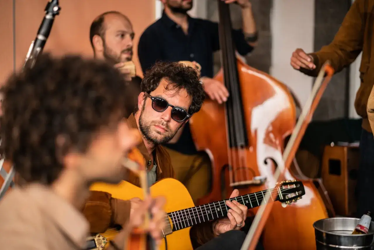 Un gruppo di musicisti suona jazz manouche. Un chitarrista con occhiali da sole è in primo piano, mentre un contrabbasso e un clarinetto sono visibili sullo sfondo.